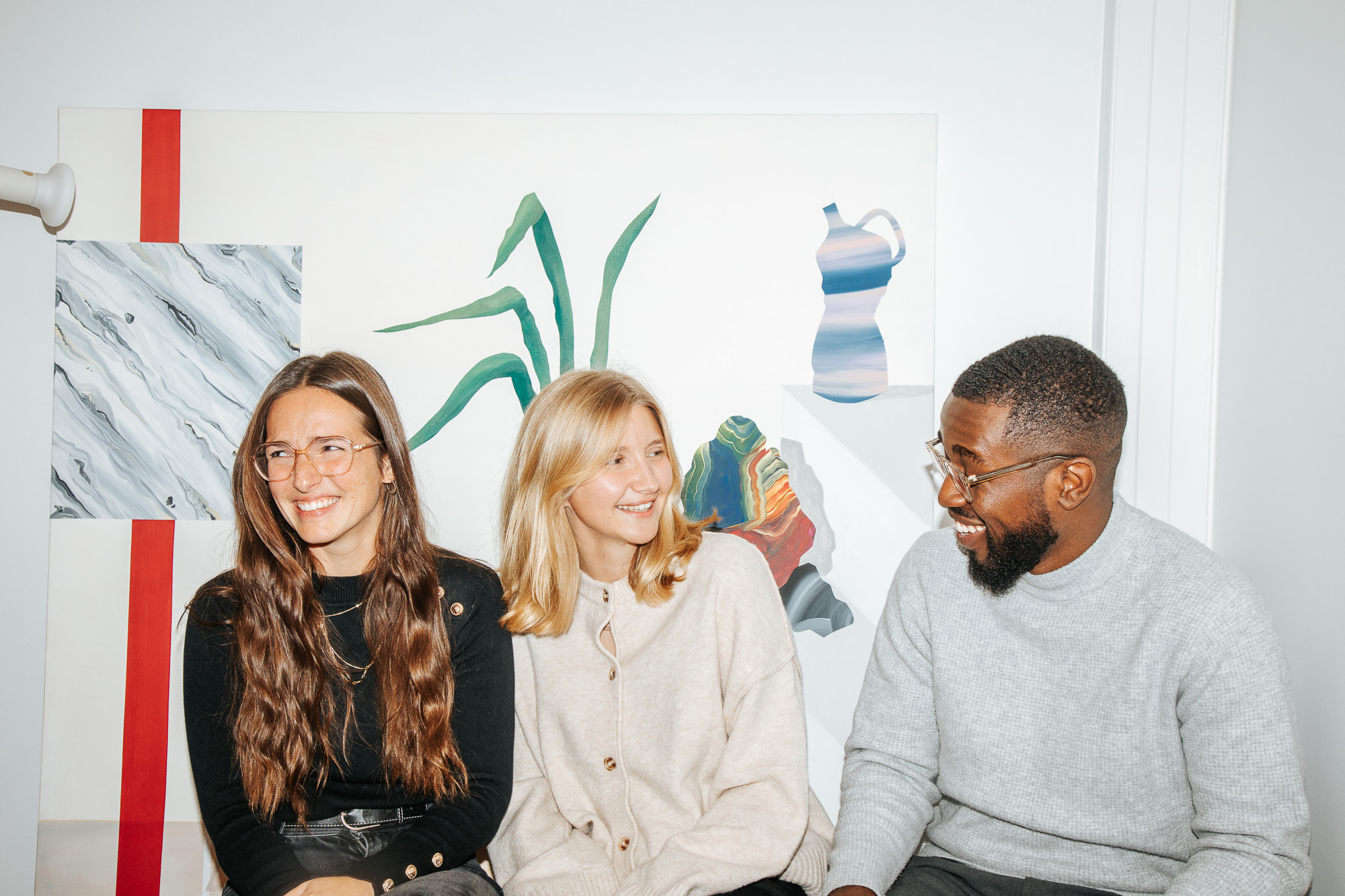 Three coworkers smiling and talking in a relaxed office workspace with artwork behind them